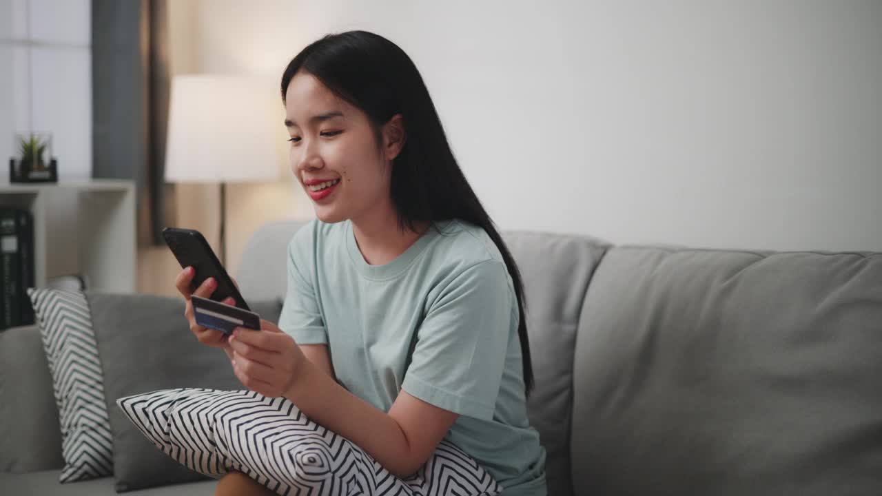 年轻女子坐在家里的沙发上用手机和信用卡网购。视频下载