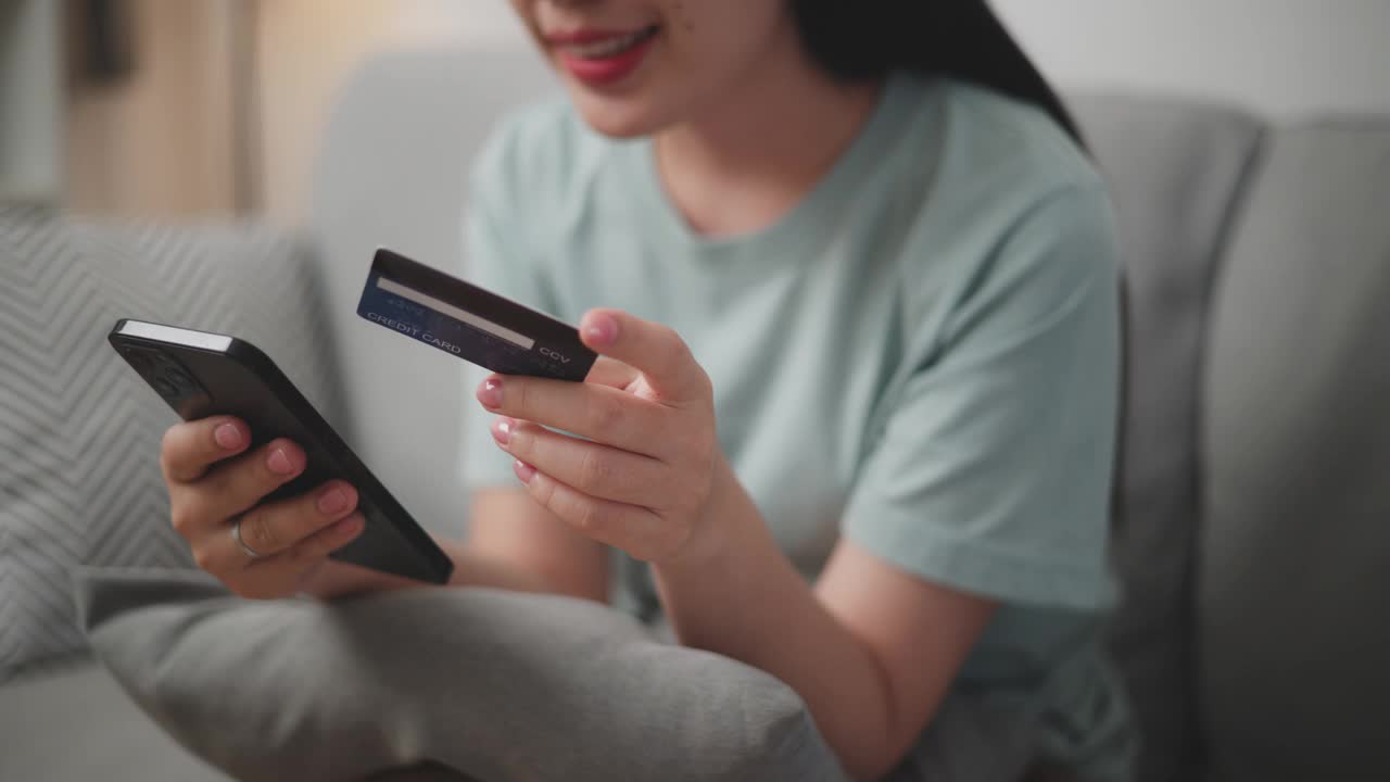 年轻女子用手机和信用卡在家里的沙发上网购。视频下载
