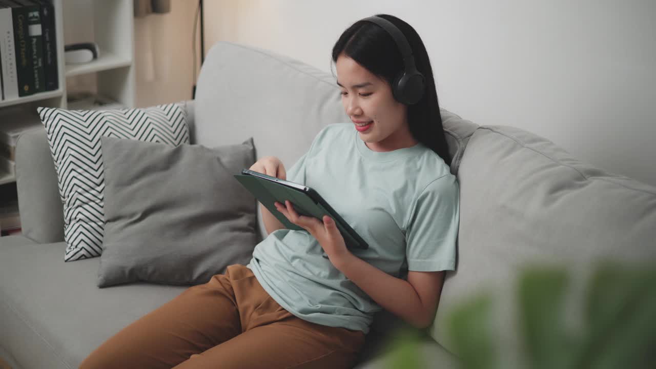 年轻女子戴着无线耳机，在家里的沙发上使用平板电脑上网购物。视频下载