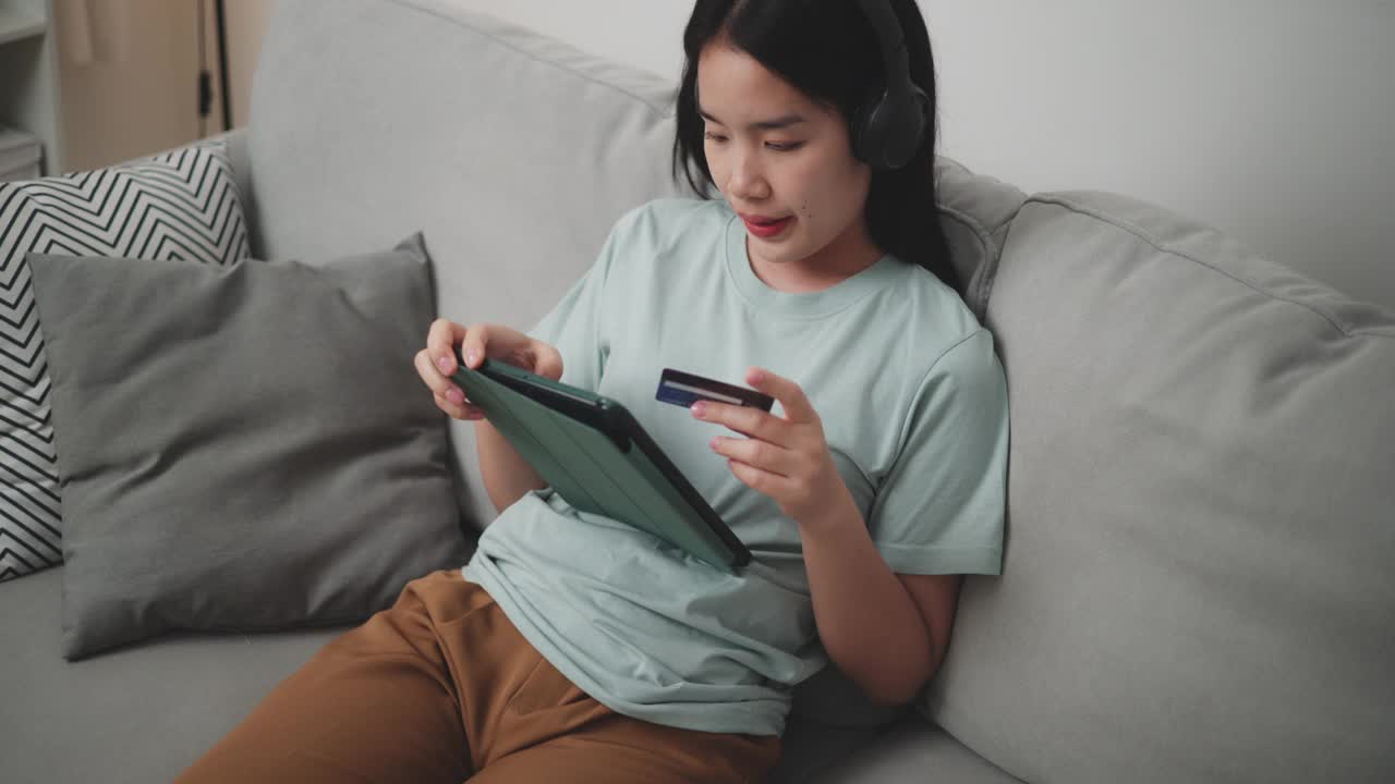 年轻女子用带无线耳机的平板电脑和信用卡坐在家里的沙发上网购。视频下载
