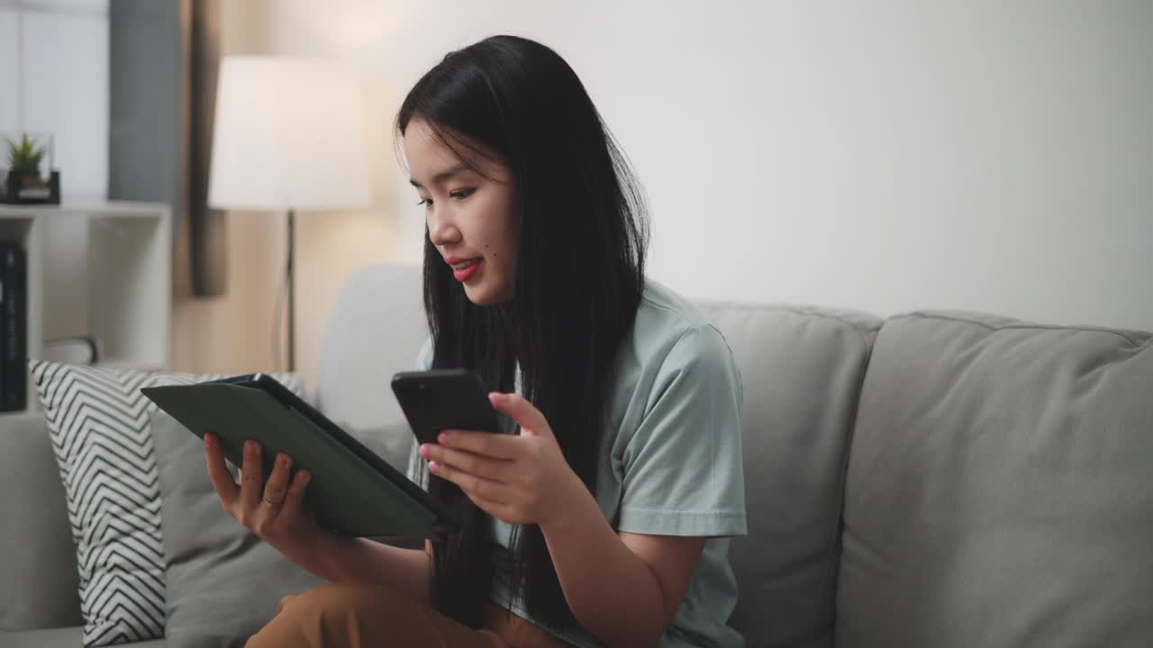 年轻女子用平板电脑和手机坐在家里的沙发上网购。视频下载