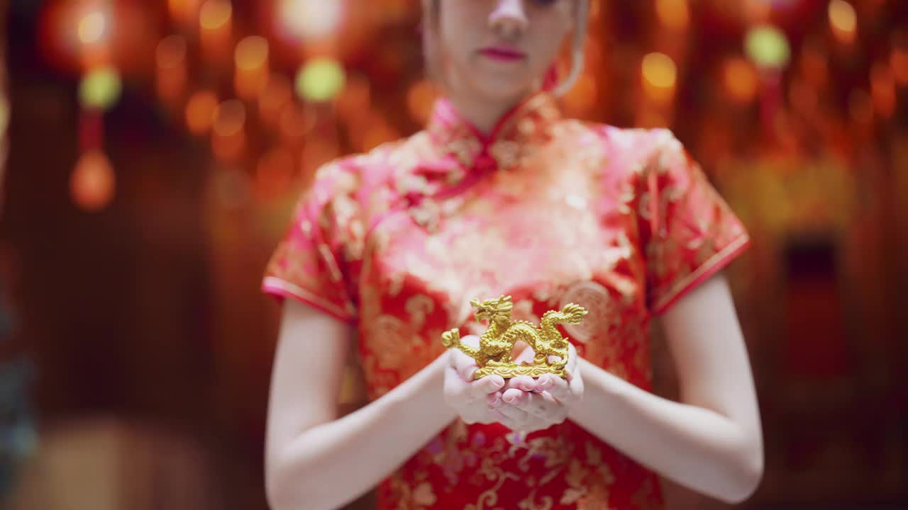 穿着红色旗袍的年轻亚洲女孩给你她手里拿着的金元宝。笑得很开心，看了看镜头。中国新年的概念。视频下载