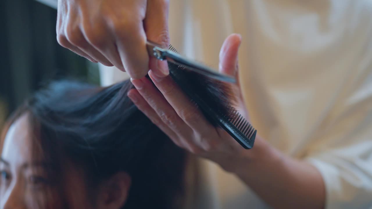 特写——专业发型师在沙龙里给女士理发，理发师用剪刀修剪黑色头发。视频下载