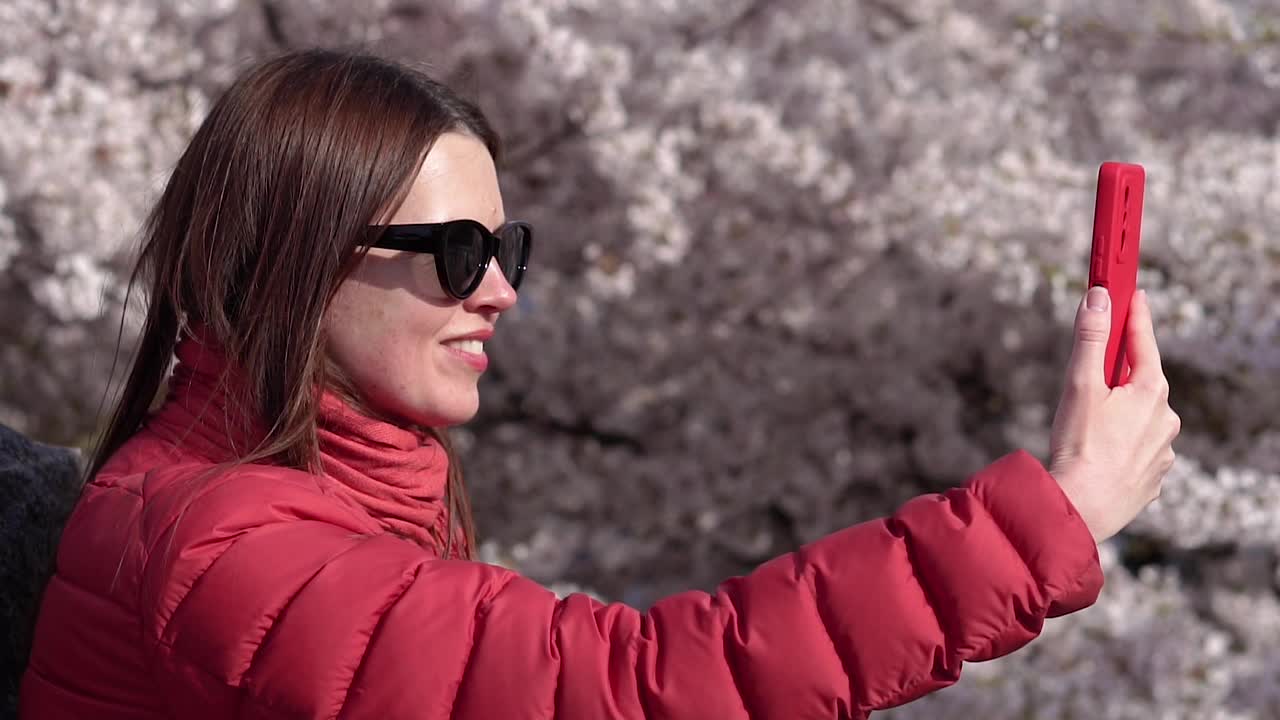 一名女子用手机给自己拍了一张照片，背景是开花的树木。视频下载