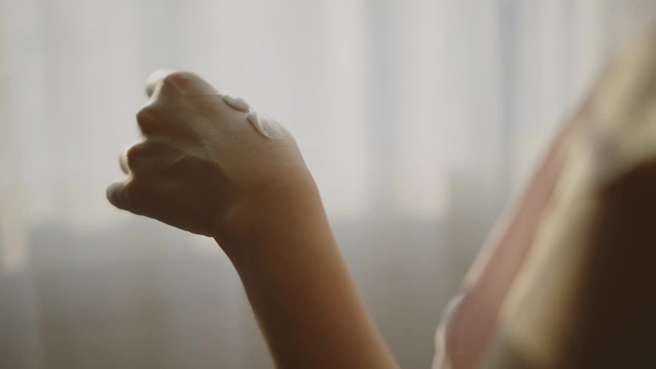 亚洲女性站在窗边给手和脸涂润肤霜的特写。视频下载