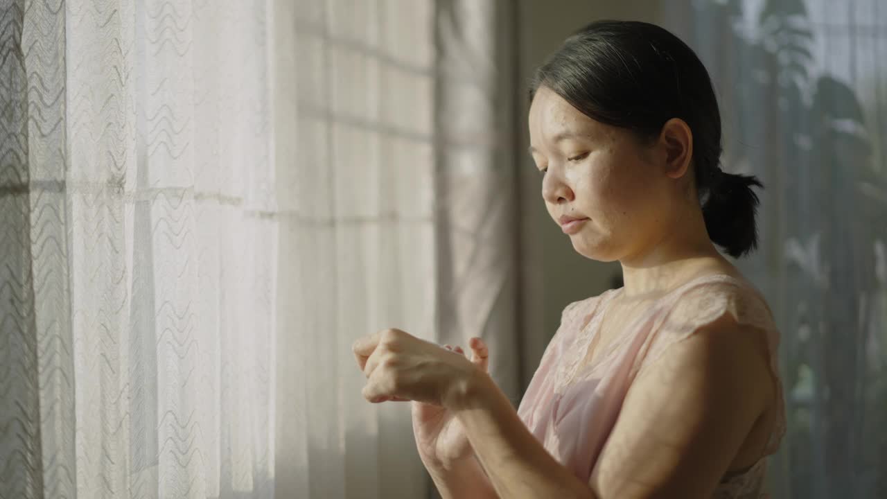 亚洲女性站在窗边，用一只手在脸颊和脖子上涂抹润肤霜。视频下载