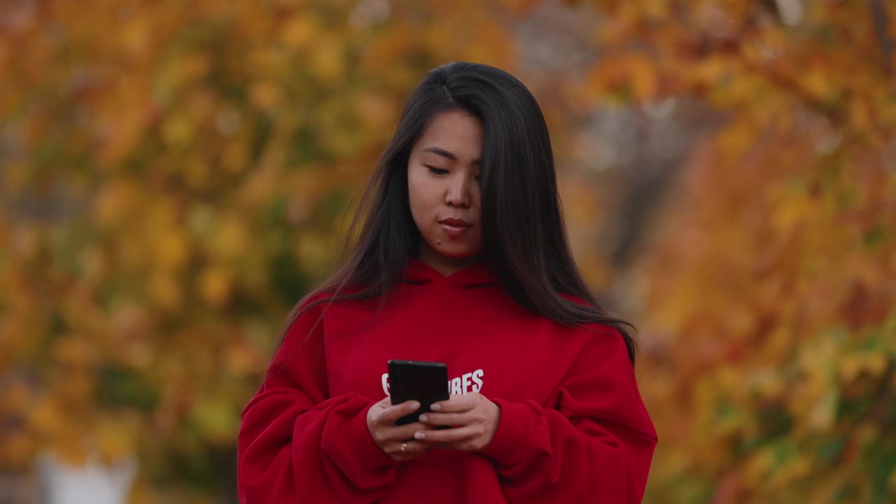 一名亚洲女子在秋天的公园里用智能手机散步视频下载
