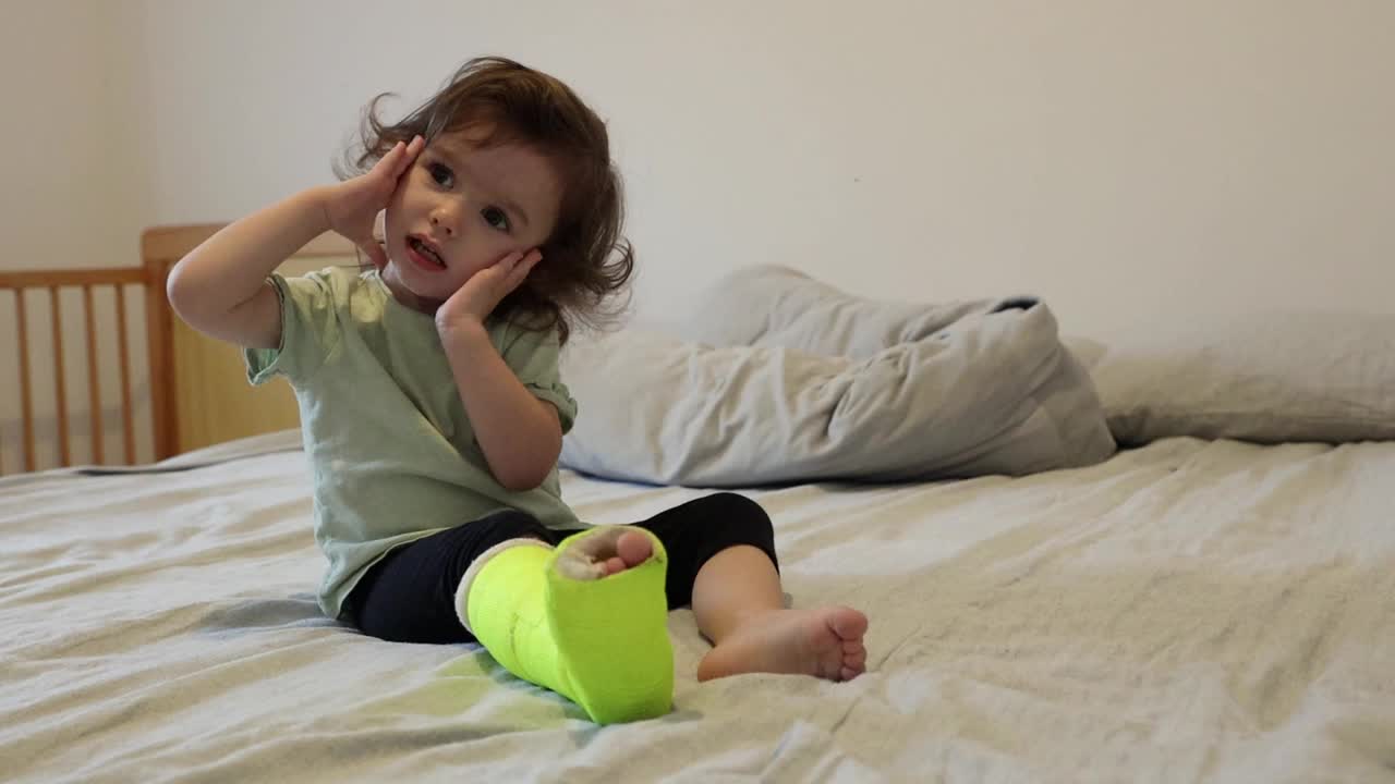 一个腿上打石膏的小女孩坐在床上调皮捣蛋。视频下载