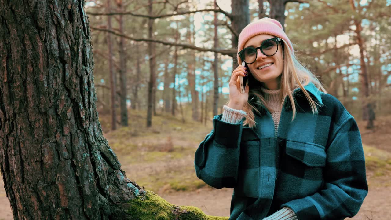 迷人的年轻女子坐在森林里的松树树干上讲手机视频下载