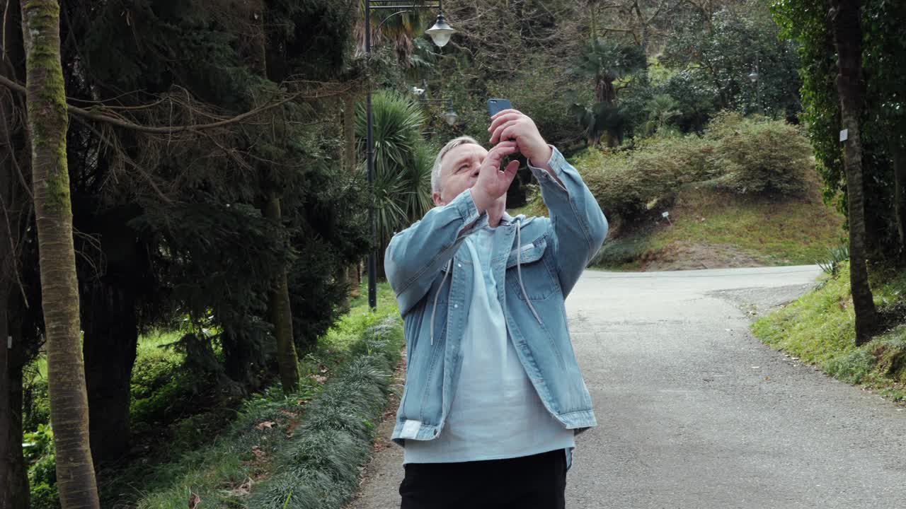 一位老人在公园里悠闲地散步，用他的智能手机捕捉周围的大自然之美视频下载