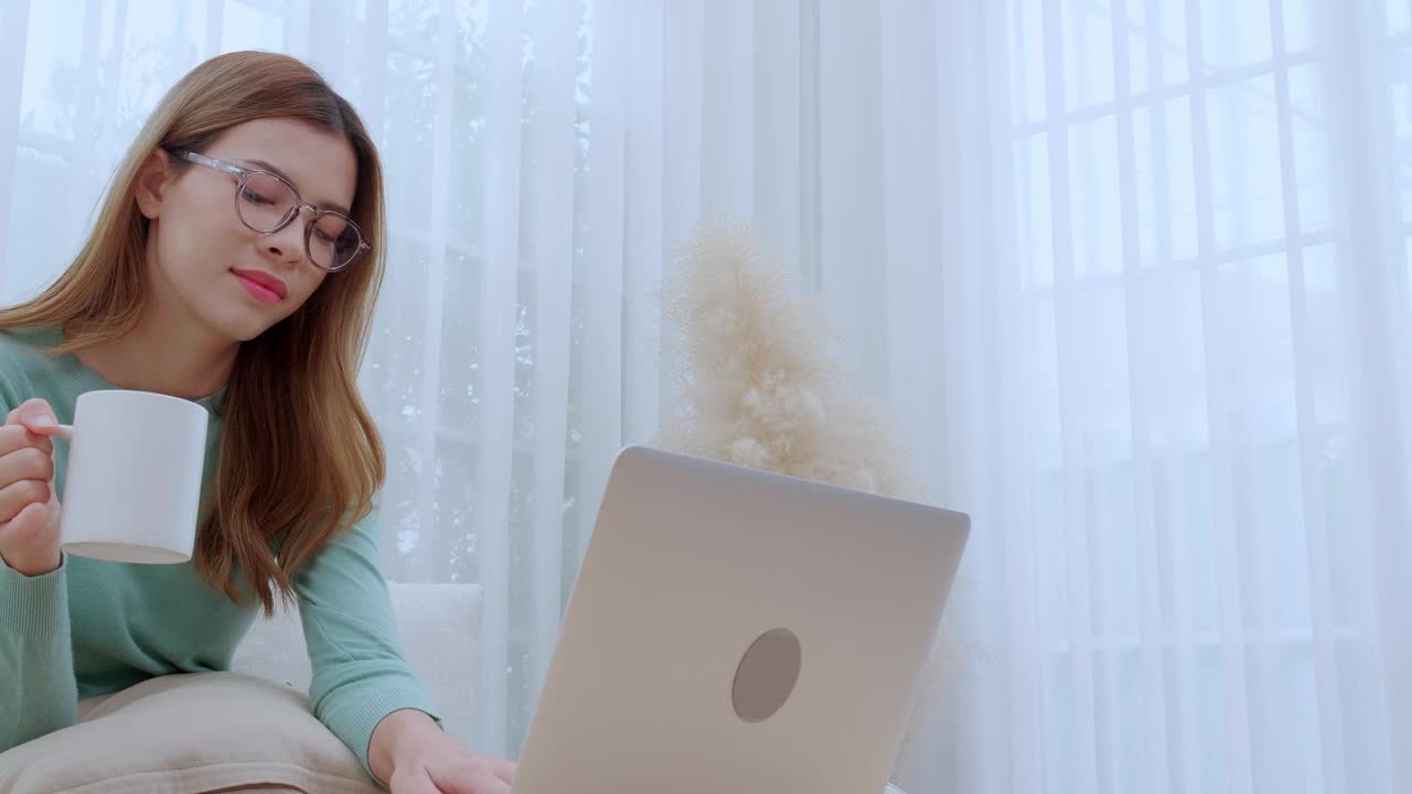 年轻的亚洲女性戴着眼镜坐在沙发上，在笔记本电脑上工作，在家里的客厅喝咖啡，女性在家工作，远程工作，自由职业，商业和数字生活。视频下载