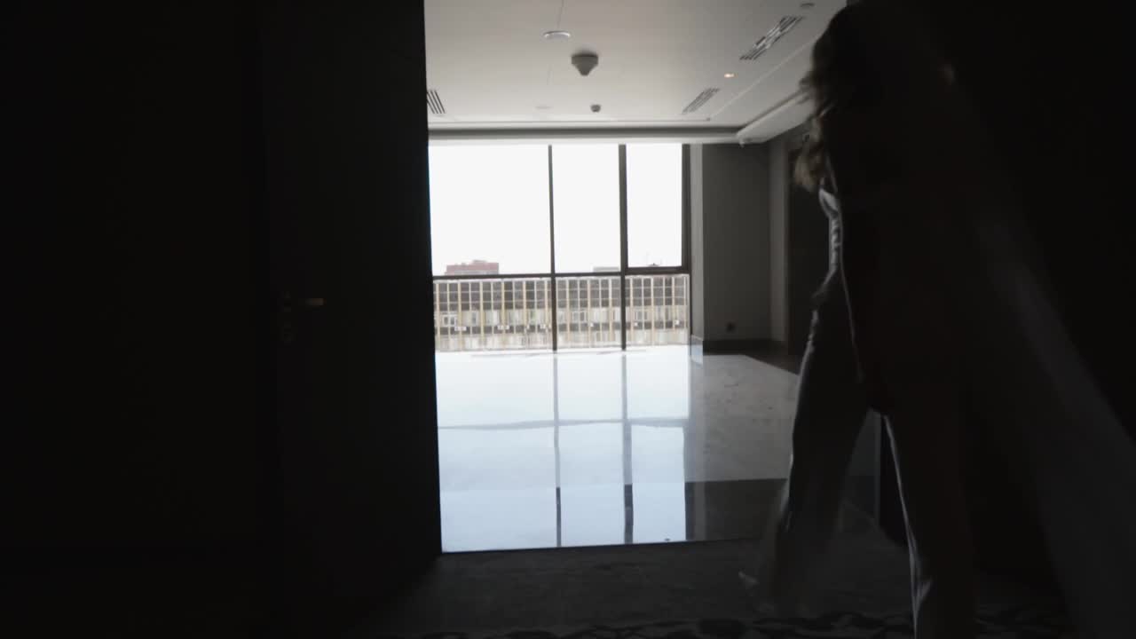 一位身穿白色长裤套装、头戴面纱的新娘沿着黑暗的走廊走向窗前。视频素材