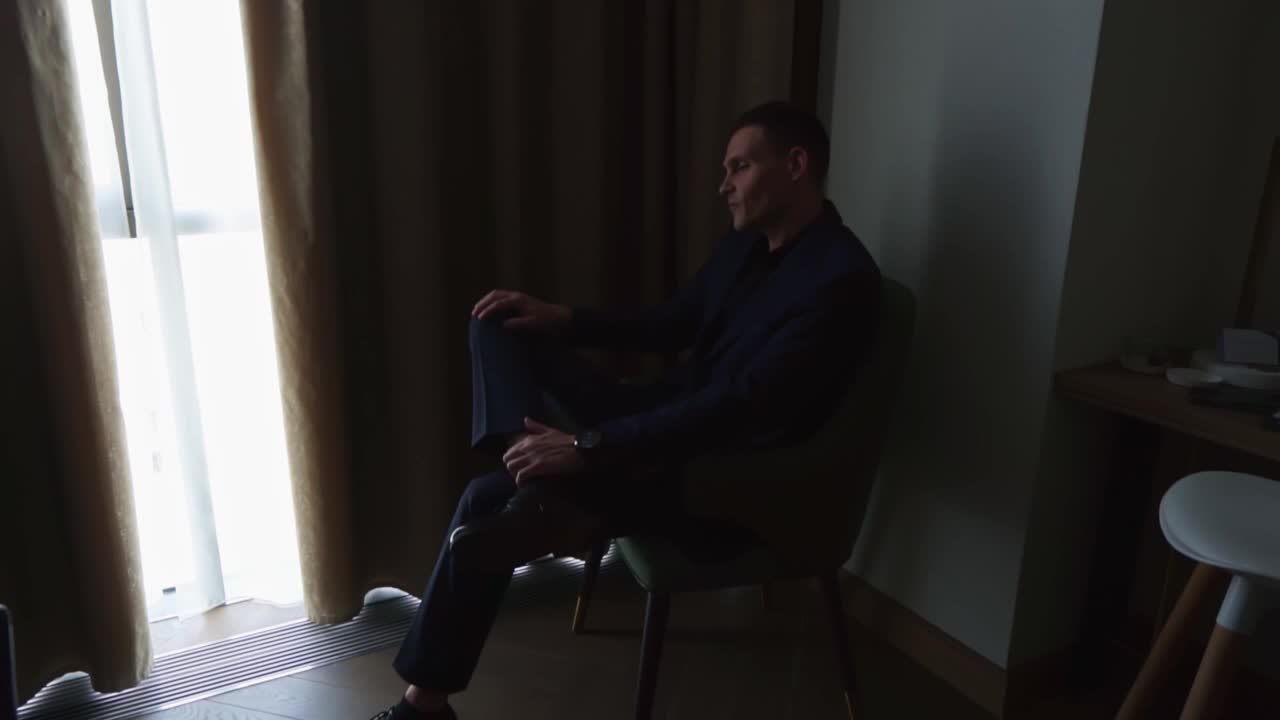一个穿着深蓝色西装的男人坐在靠窗的椅子上的扶手椅上。视频下载