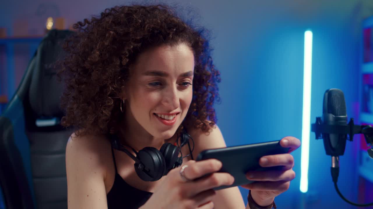 快乐的女人女孩玩家戴着耳机竞争玩视频游戏在线与智能手机彩色霓虹灯在家里的客厅。电子竞技直播游戏在线，居家隔离活动概念。视频下载