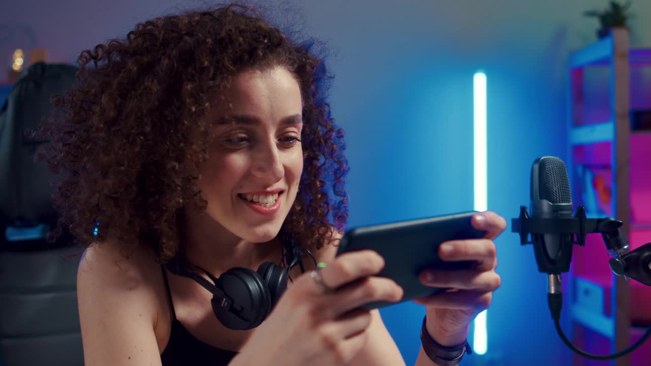 快乐的女人女孩玩家戴着耳机竞争玩视频游戏在线与智能手机彩色霓虹灯在家里的客厅。电子竞技直播游戏在线，居家隔离活动概念。视频下载