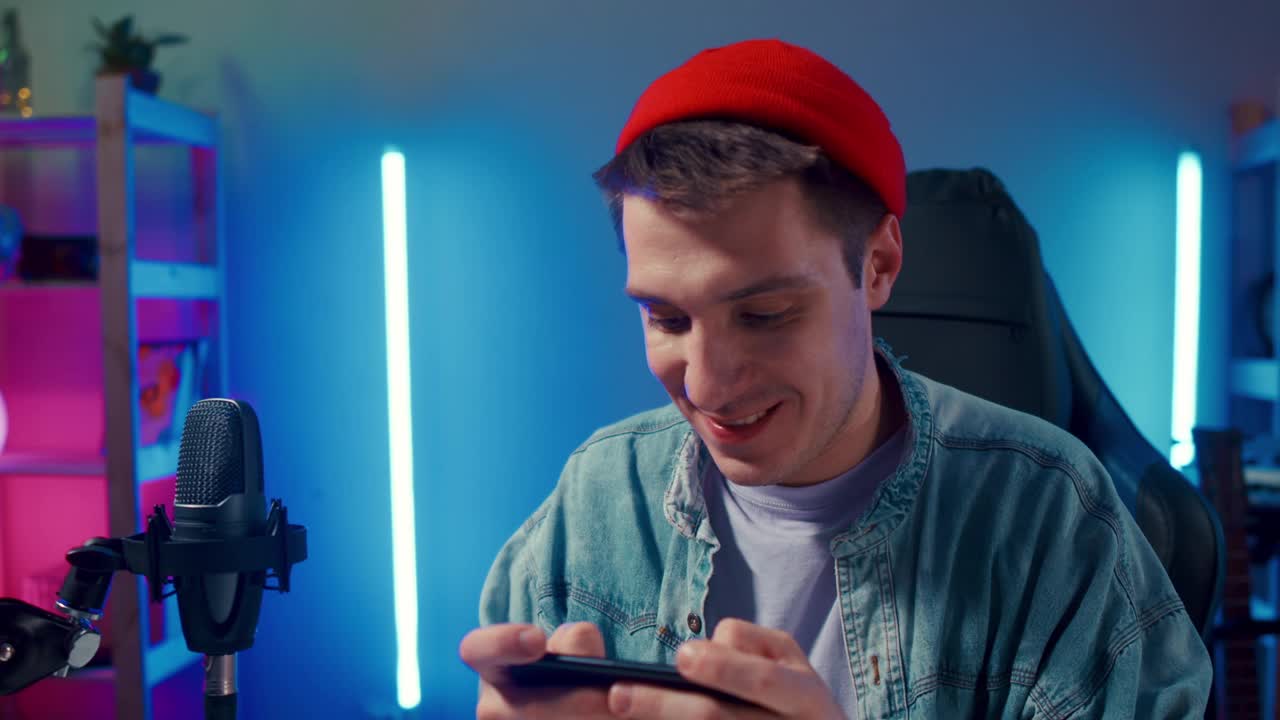 快乐的男人玩家戴着红帽子比赛玩视频游戏在线与智能手机彩色霓虹灯在客厅晚上现代的房子。在线电子竞技直播游戏，居家隔离活动。视频下载