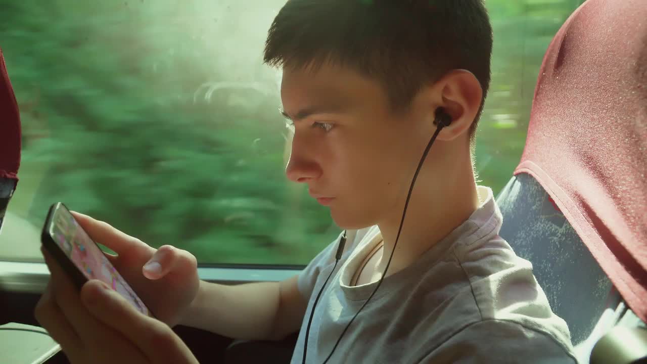 关闭了。一个十几岁的男孩坐在公共交通穿梭巴士的窗口边玩网络游戏边玩智能手机。在火车上通过有线耳机写信息听音乐视频素材