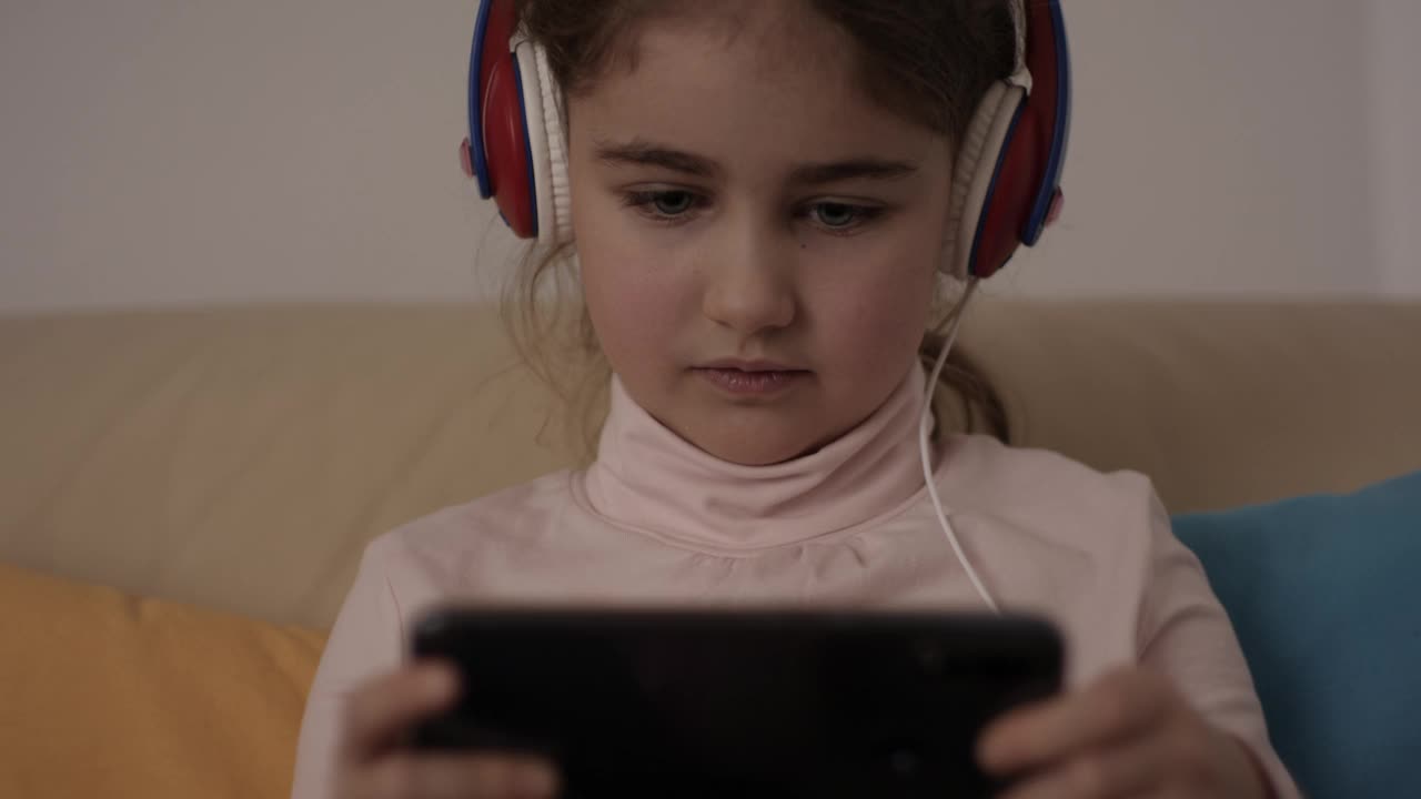 孩子玩手机玩得很开心。小女孩戴着耳机在手机上玩视频游戏。青少年用智能手机玩电子游戏对儿童心理健康视力的危害视频下载