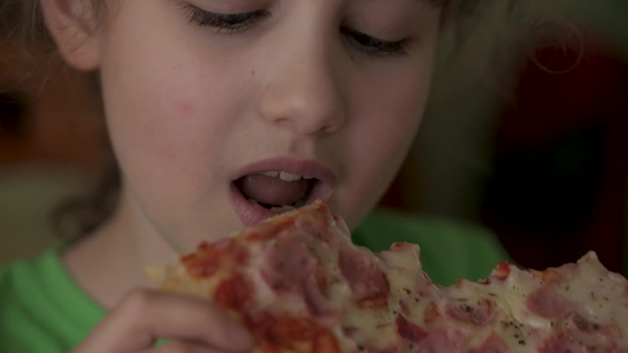 饥饿的孩子咬了一口披萨。吃披萨。饥饿的小男孩坐在家里厨房的餐桌上吃着美味的意大利披萨。不健康的营养快餐。一个吃午饭的孩子的肖像。视频下载