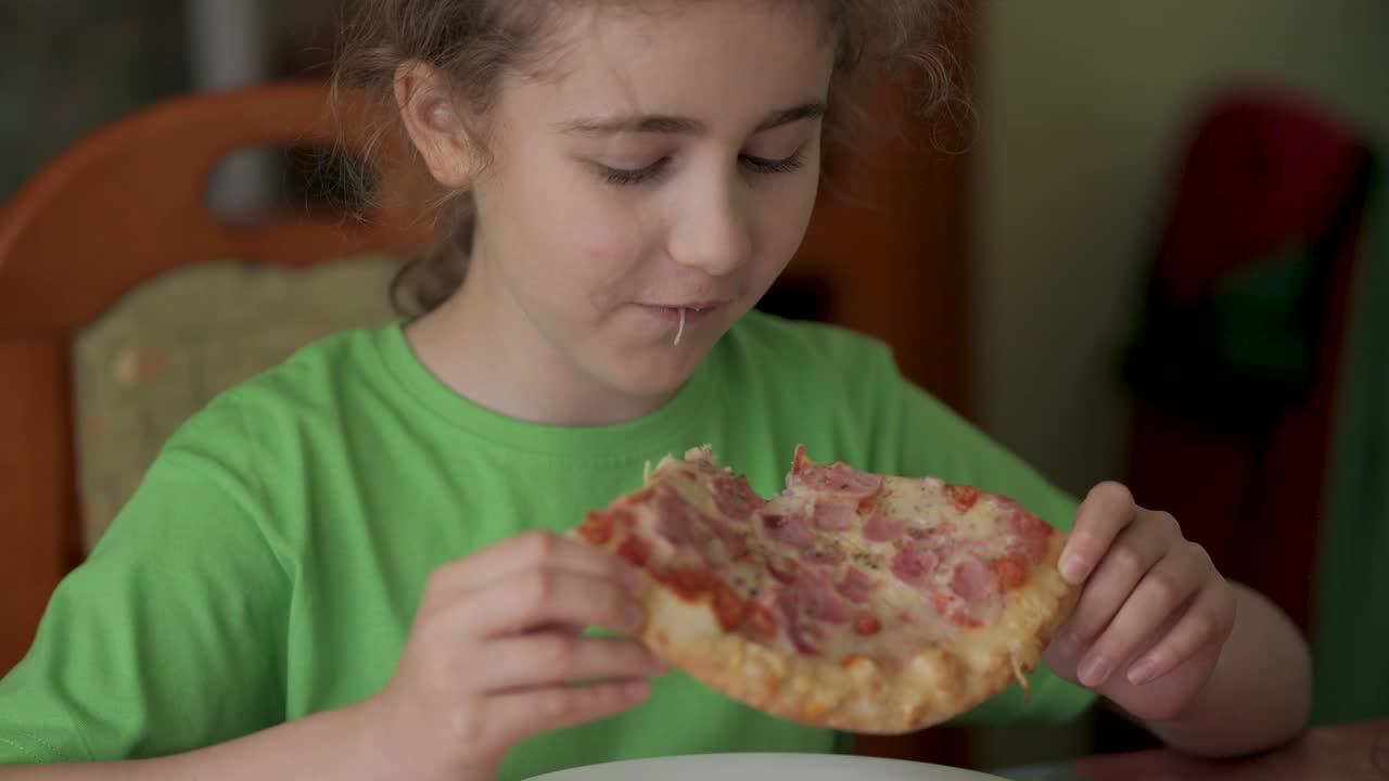饥饿的孩子咬了一口披萨。吃披萨。饥饿的小男孩坐在家里厨房的餐桌上吃着美味的意大利披萨。不健康的营养快餐。一个吃午饭的孩子的肖像。视频下载