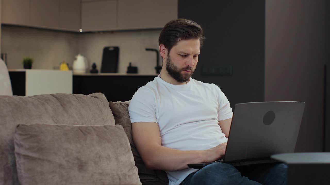 随意的男人坐在客厅的沙发上使用便携式电脑。男人在家工作，网上购物。现代科技、互联网和人本理念。视频下载