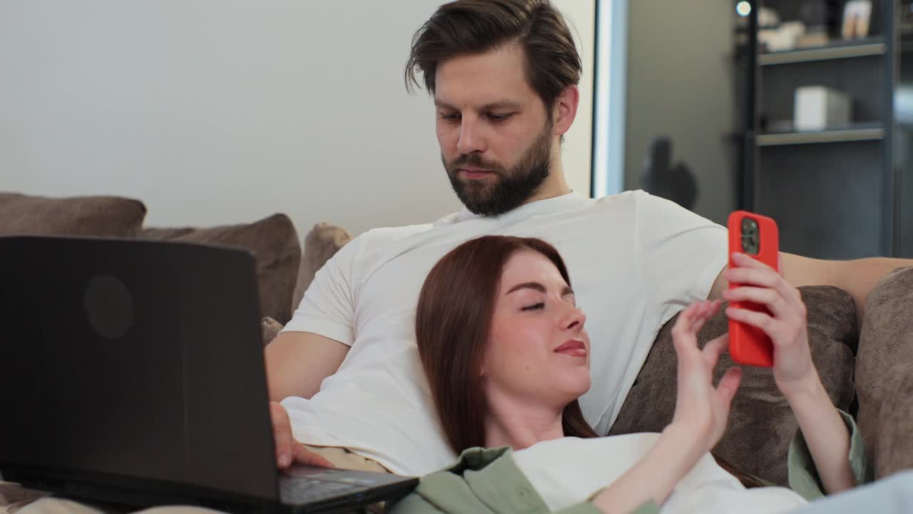 一对夫妇在沙发上放松，男人在笔记本电脑上工作，女人跪在男人身上，在家用手机刷社交媒体。远程工作的概念。视频下载
