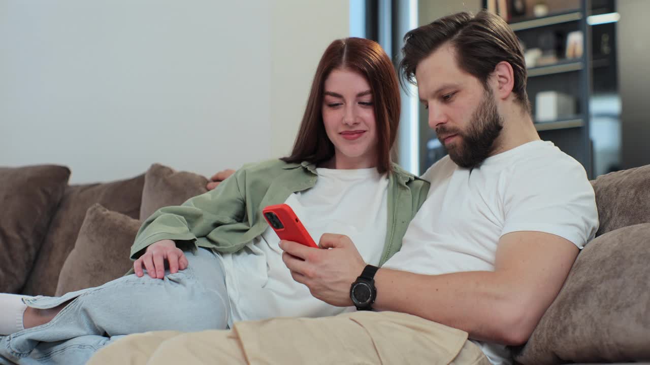 幸福的已婚夫妇男女在家里的沙发上使用智能手机。使用手机应用程序在网上商店购物，订购送货。视频下载