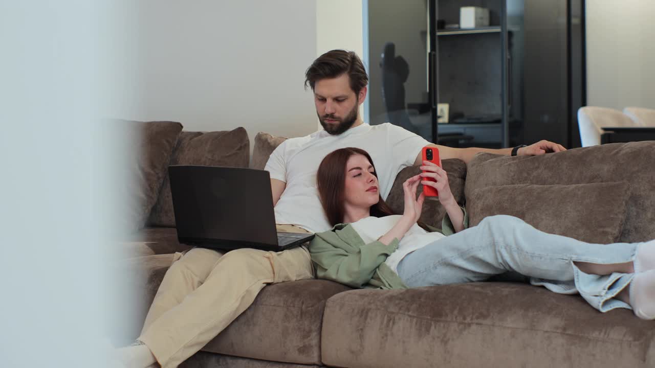 幸福的家庭夫妇在沙发上放松，男人在笔记本电脑上工作，女人，妻子，跪在男人身上，在家里用手机刷社交媒体。远程工作的概念。视频下载