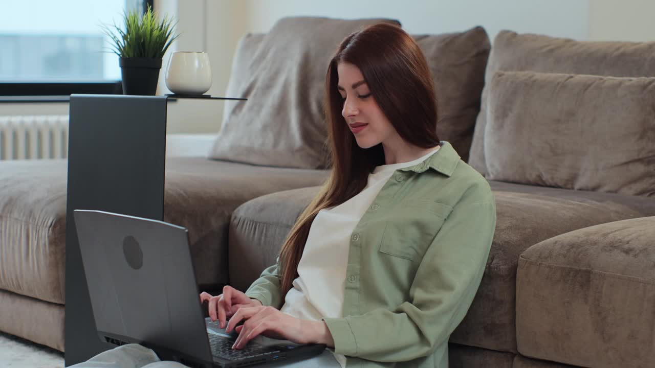 年轻女子坐在公寓的客厅地板上使用手提电脑。女人在家工作，在网上购物。现代科技、互联网和人本理念。视频下载
