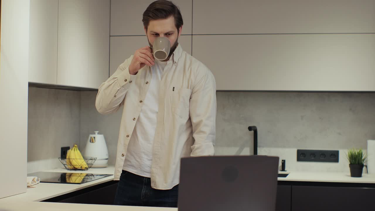 留着胡子的年轻男子在自家厨房用笔记本电脑。男性喝咖啡。在家工作。科技与人的理念。视频下载