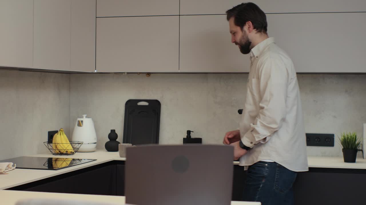 留着胡子的年轻男子在自家厨房用笔记本电脑。男性喝咖啡。在家工作。科技与人的理念。视频下载