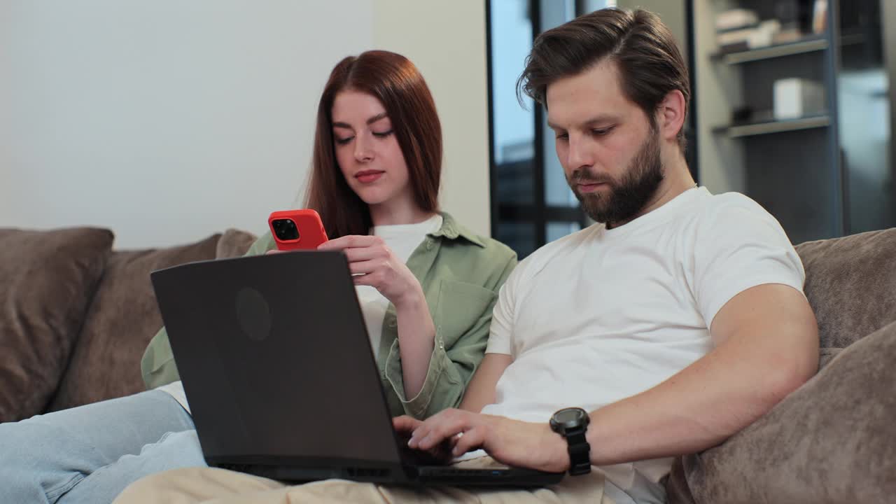 这对夫妇在公寓客厅的沙发上休息，一边使用笔记本电脑和智能手机。男人和女人在聊天，在网上购物，在网上订购产品。视频下载
