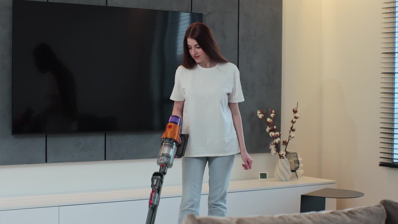 年轻女子用吸尘器打扫家里客厅的地板。使用无线吸尘器清洁家居。智能清洁技术。视频下载