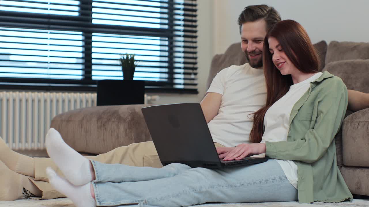 一对夫妇在他们公寓的地板上使用笔记本电脑。男人和女人在聊天，在网上购物，在网上订购产品。视频下载