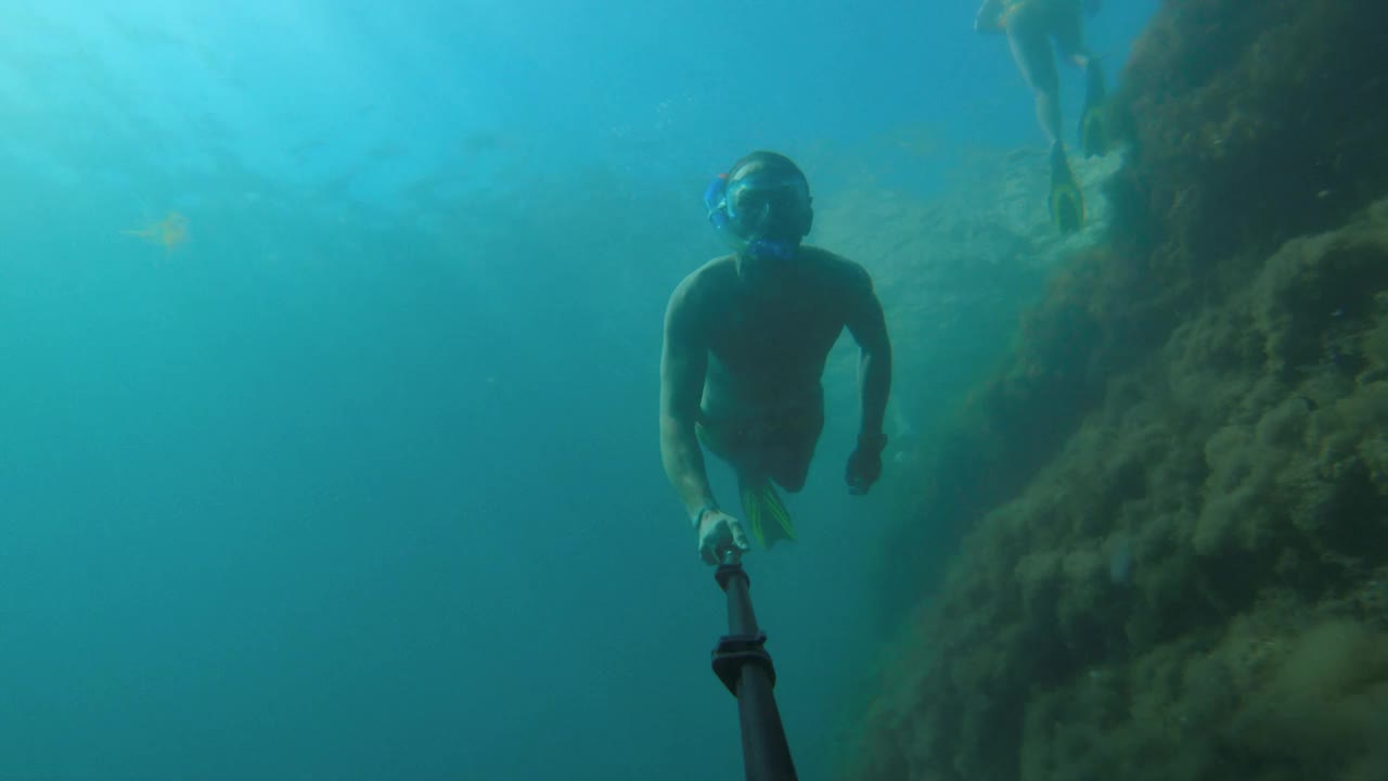 一名白人男子戴着潜水面罩，穿着脚蹼，拿着一根长自拍杆上的运动相机在水下潜水和游泳。浮潜年成视频下载