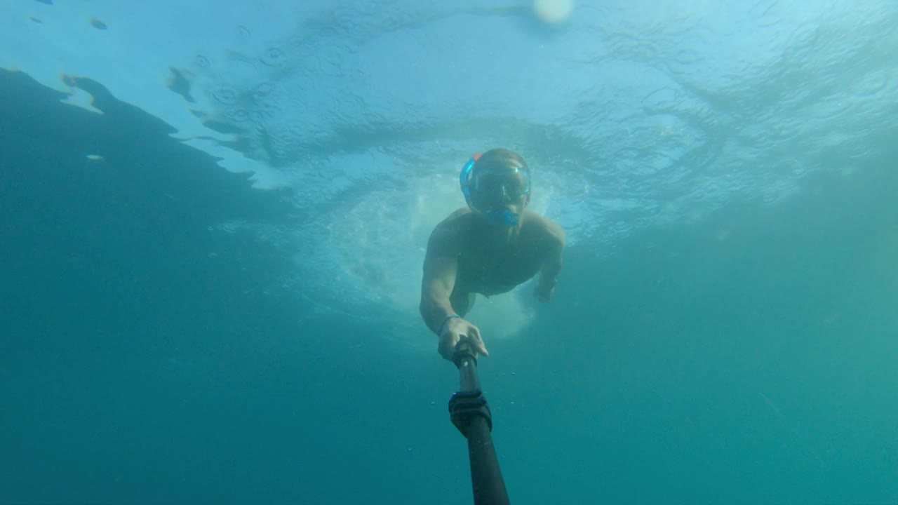 一名白人男子戴着潜水面罩，穿着脚蹼，拿着一根长自拍杆上的运动相机在水下潜水和游泳。浮潜年成视频素材