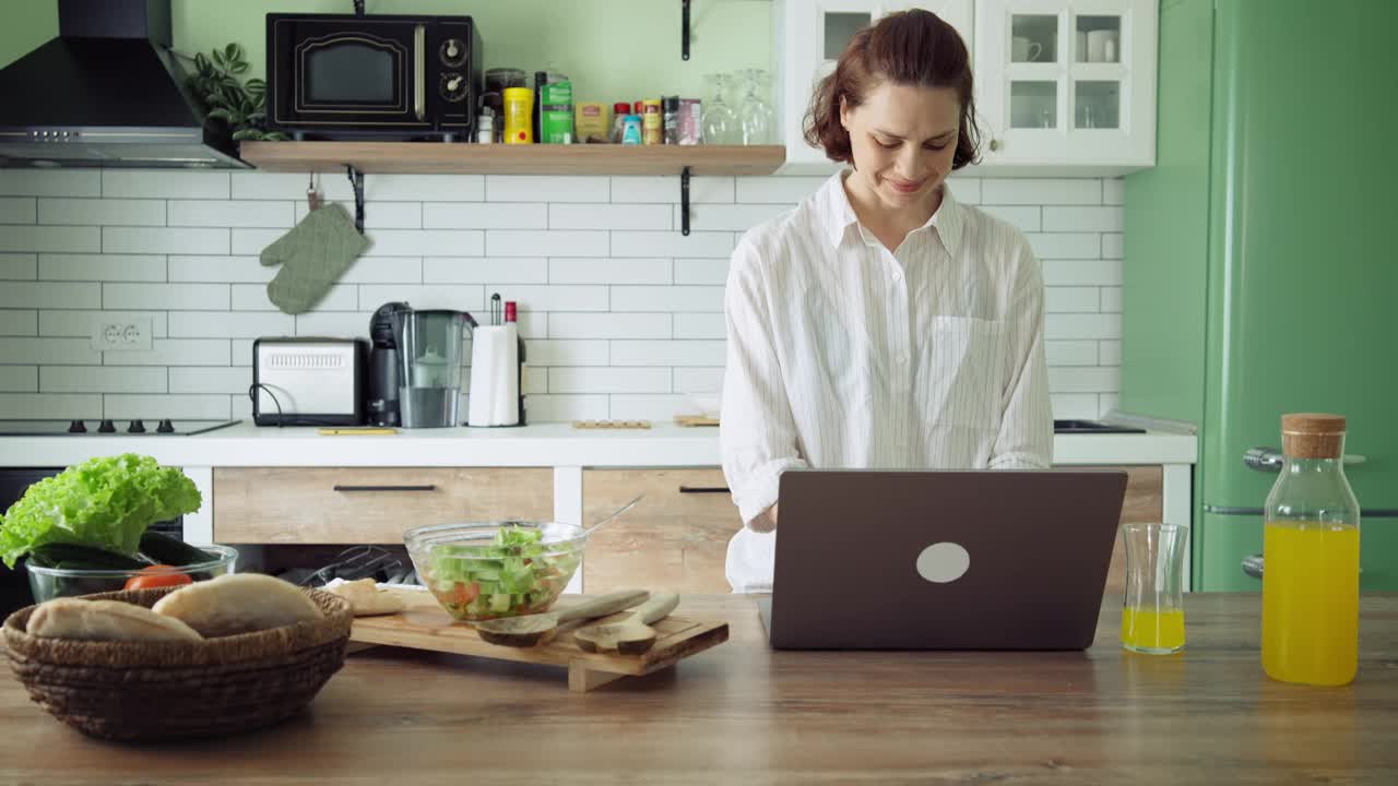 一位年轻的白人女性站在明亮的厨房里使用笔记本电脑。视频下载