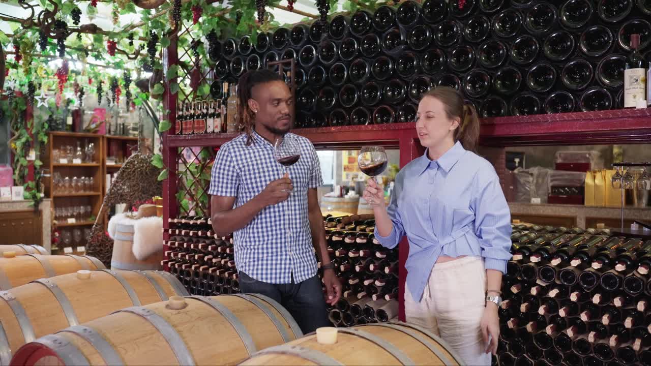 两位专业的侍酒师，中年非洲黑人男性和白人女性在一个迷人的老酒窖，享受聊天和品尝优质的红葡萄酒在葡萄酒工厂。视频下载