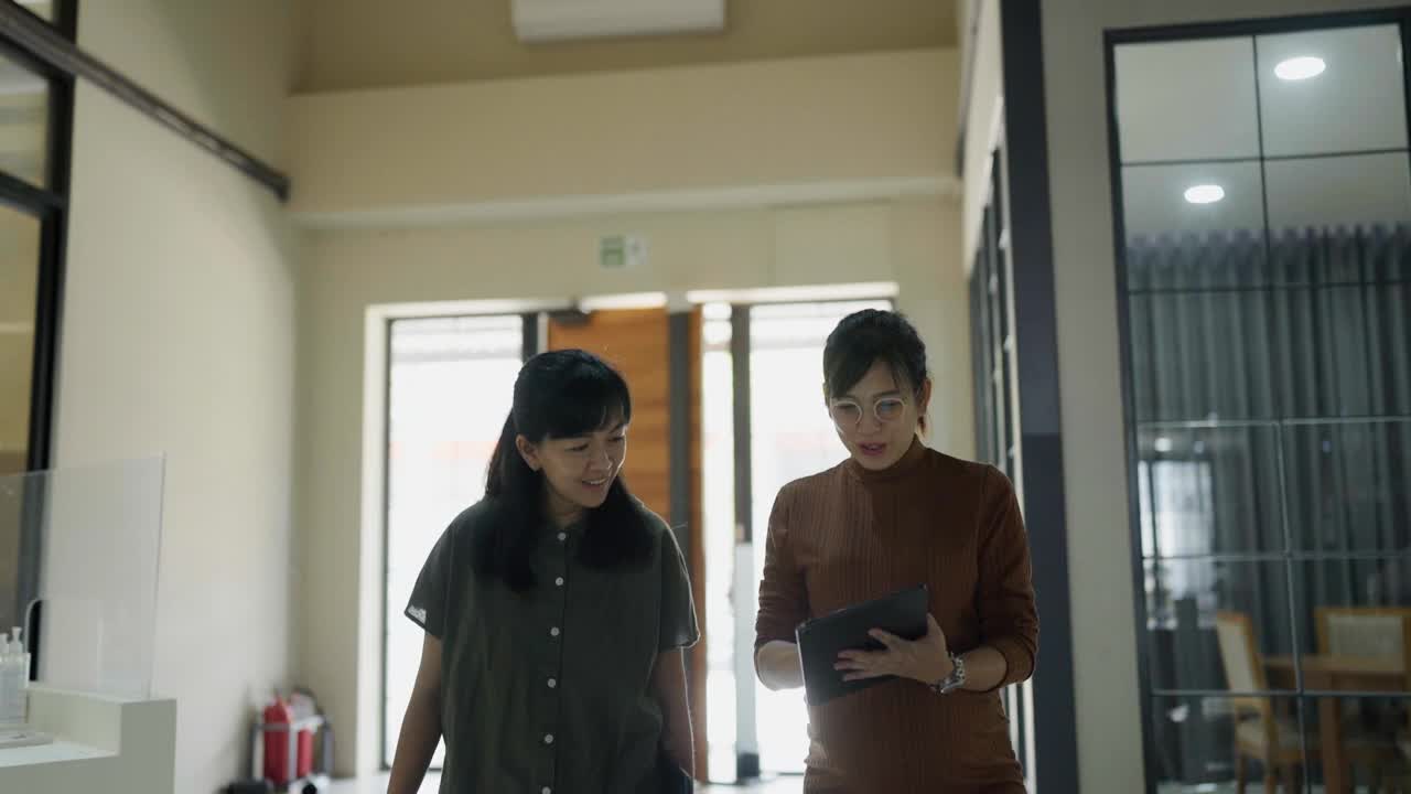怀孕的秘书走在办公室走廊上，拿着数码平板电脑向女商人做会议报告的正面照片。视频下载