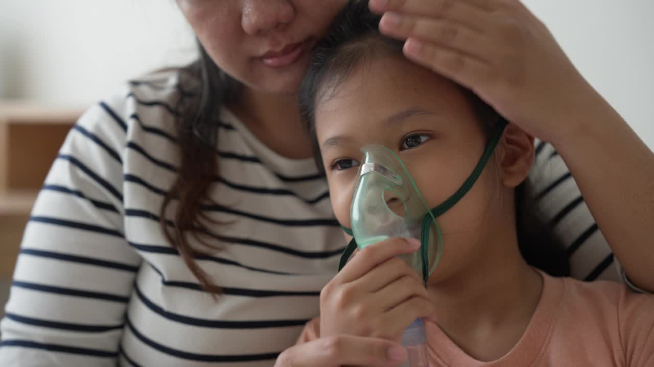 一位亚洲母亲在家照顾生病的女儿，她坐在床上接受喷雾器治疗。哮喘的治疗视频下载