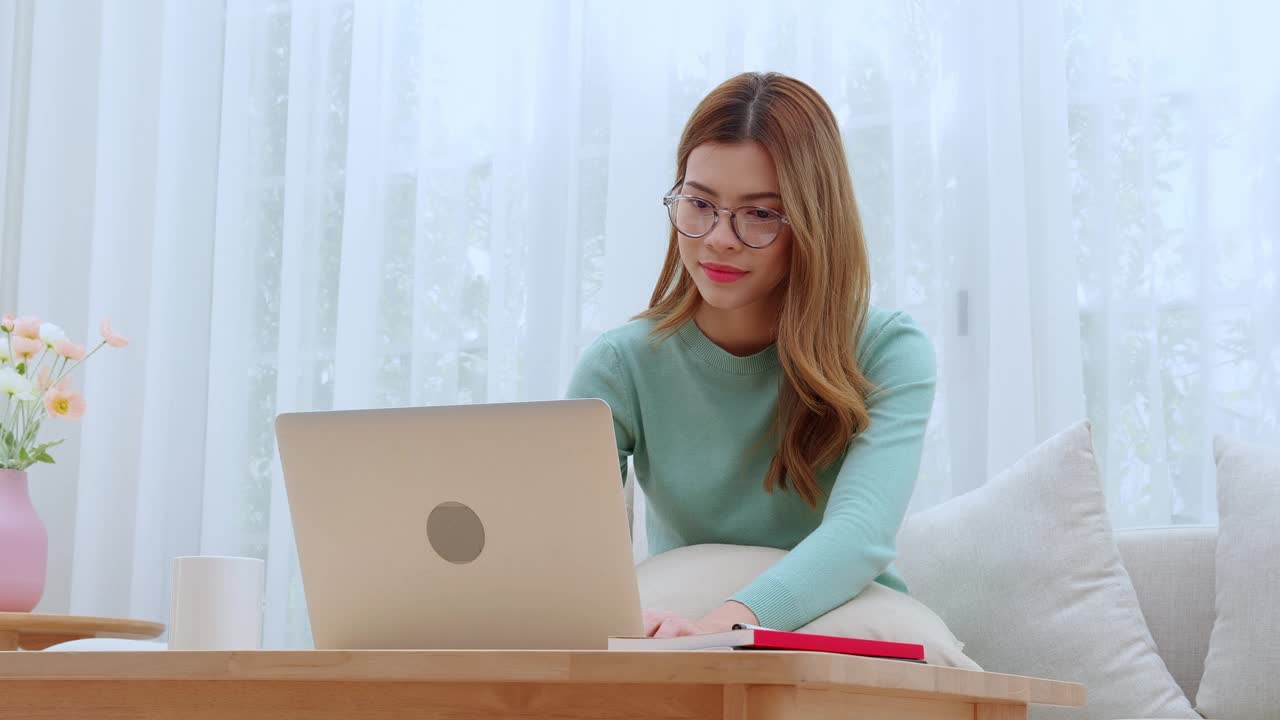 年轻的亚洲女性戴着眼镜坐在沙发上在家里客厅的笔记本电脑上工作，女性在家工作，远程办公，自由职业者在工作空间，商业和数字生活。视频下载