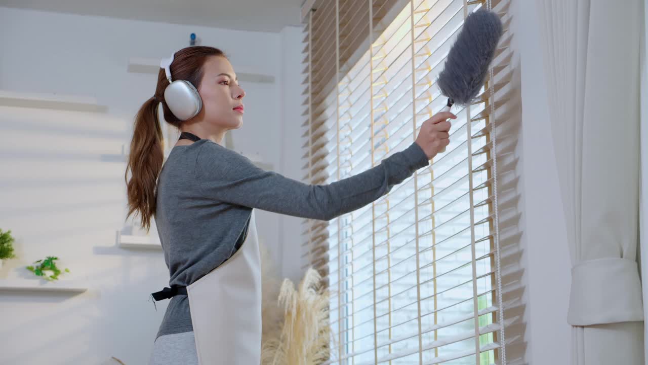 年轻的亚洲女性在明亮整洁的客厅里一边打扫一边戴着耳机听音乐，女佣在家里打扫一边听音乐和唱歌，生活方式概念。视频下载
