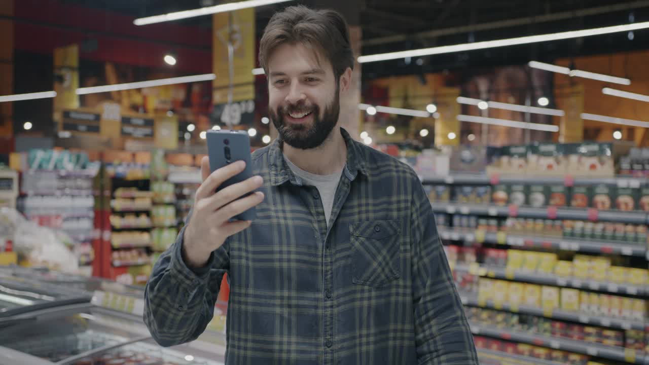 年轻人在超市用智能手机进行在线视频通话，展示食品，讨论购买视频下载