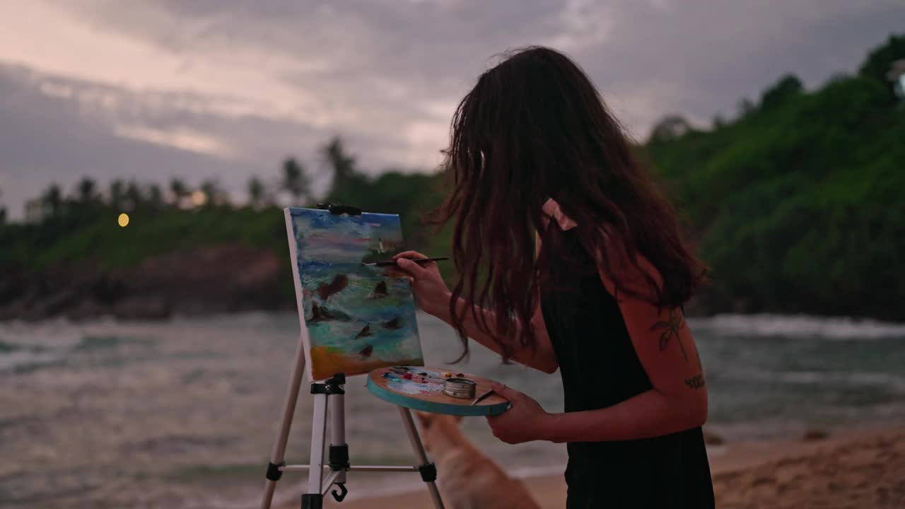 一位女艺术家在黄昏的沙滩上用画布描绘汹涌的大海。海浪撞击，喜怒无常的天空，手里拿着画笔，专注于创作暴风雨般的海洋艺术品，灵感奔涌。Slowmo视频下载