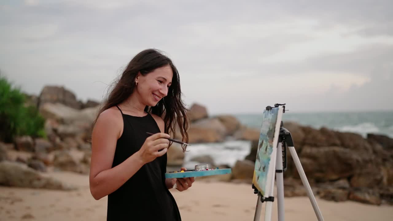 在岩石岸边的帆布上，黑色连衣裙的艺术家描绘黄昏的大海。风吹发，浪撞，创意心情。女人微笑着，拿着画笔，在调色板上混合颜色，捕捉海岸之美。Slowmo视频下载