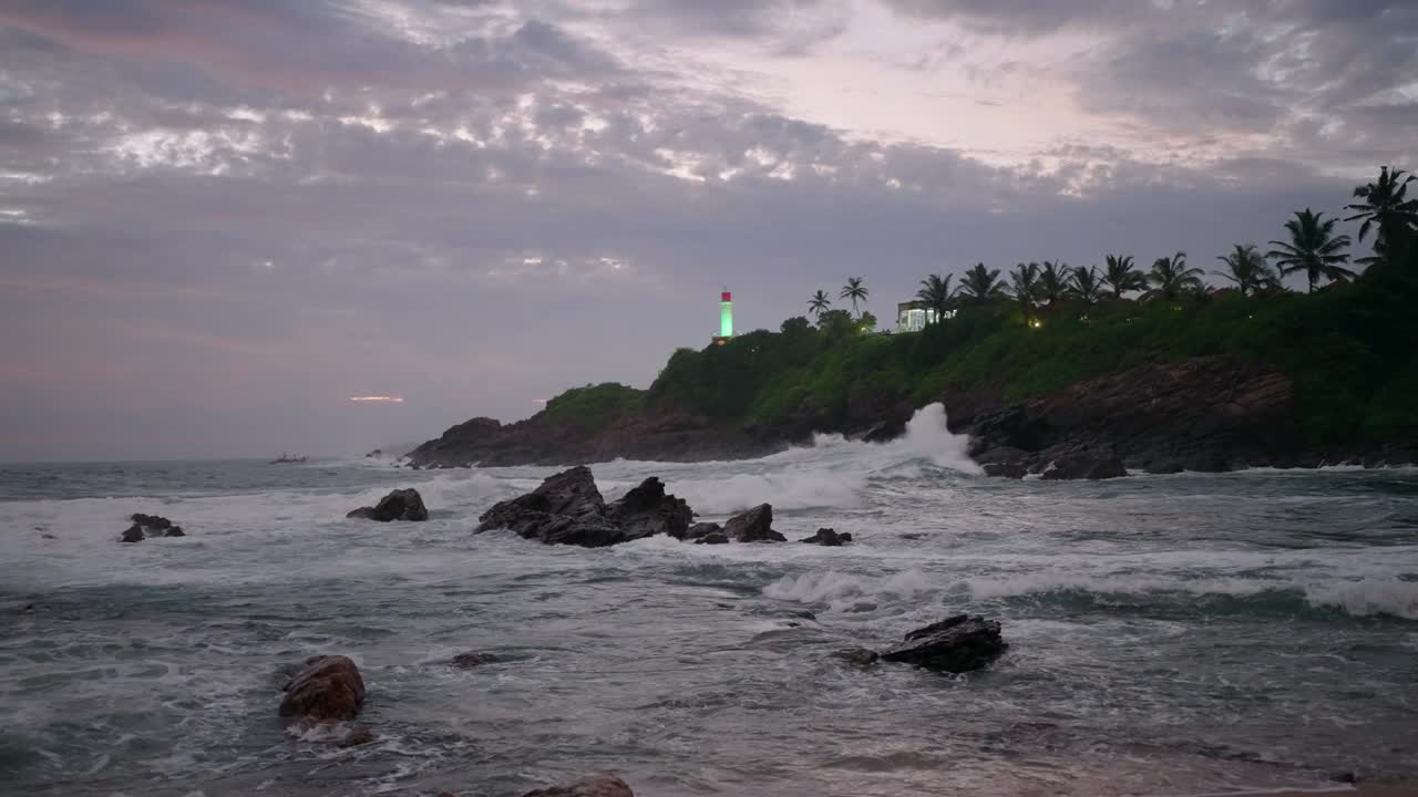 黄昏时分，波涛汹涌，布满岩石的海岸。黄昏时海浪拍打着石头。热带景观中的灯塔是绿色的。日落后大海上紫色的天空。自然景观与水的运动。Slowmo视频下载