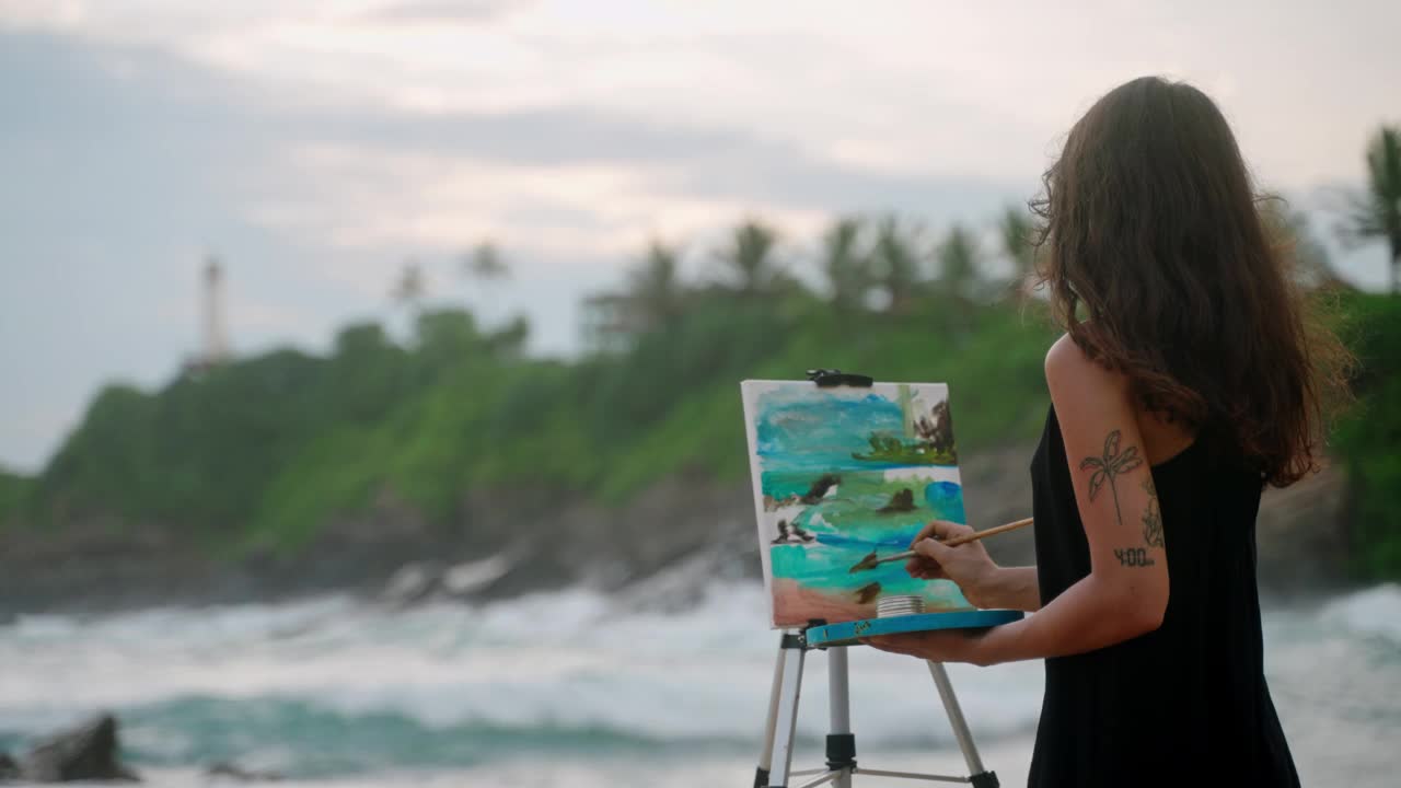 一位女艺术家在狂风暴雨的海滩上，用画布描绘出色彩斑斓的海景。纹身可见的手臂上增加了艺术品，灯塔背景上充满活力的细节。艺术爱好，海滨自然视频下载