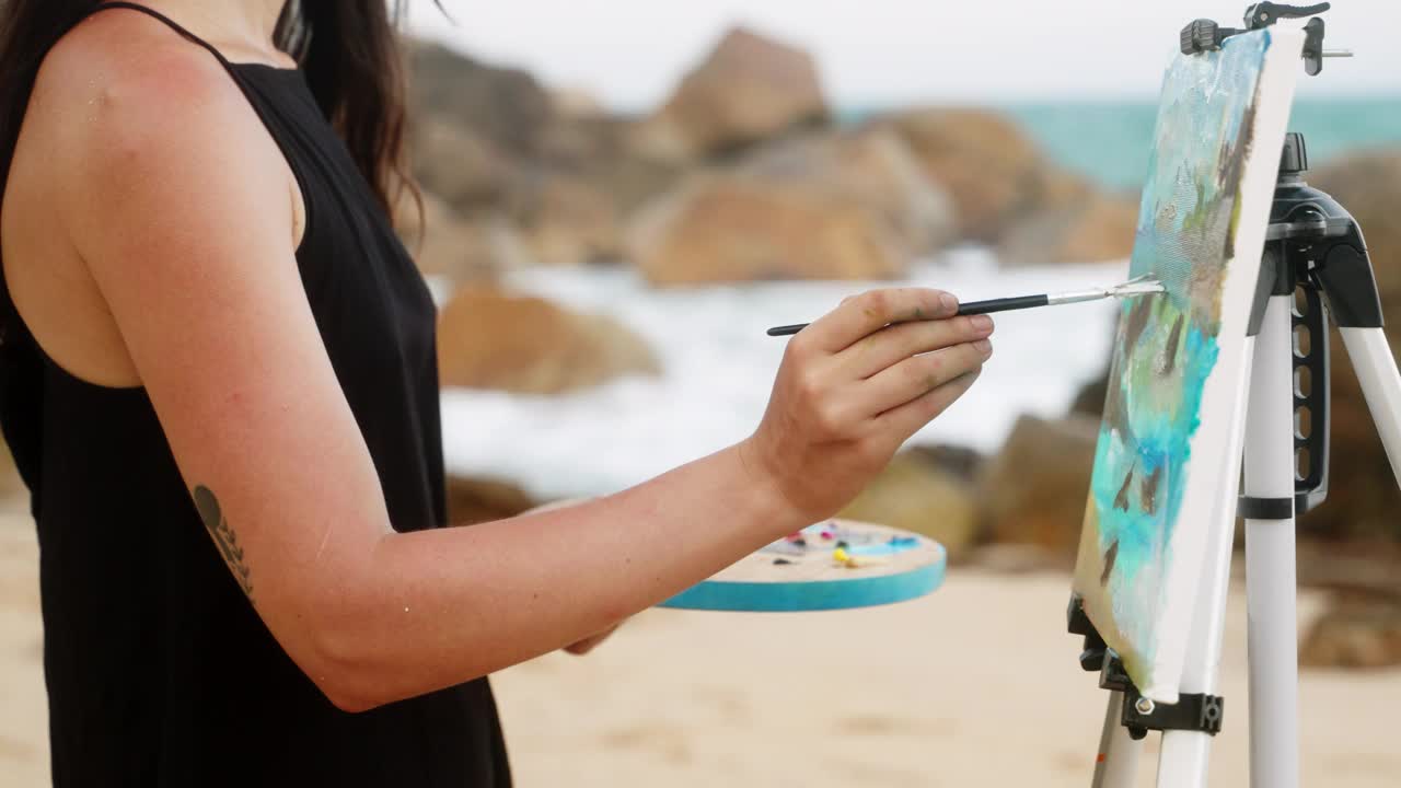 艺术家在沙滩海岸线的画布上描绘汹涌的海浪。创意女性用画笔、调色板描绘海洋能量。业余爱好者，专注于海边的艺术创作。大自然激发了户外艺术的灵感。视频下载
