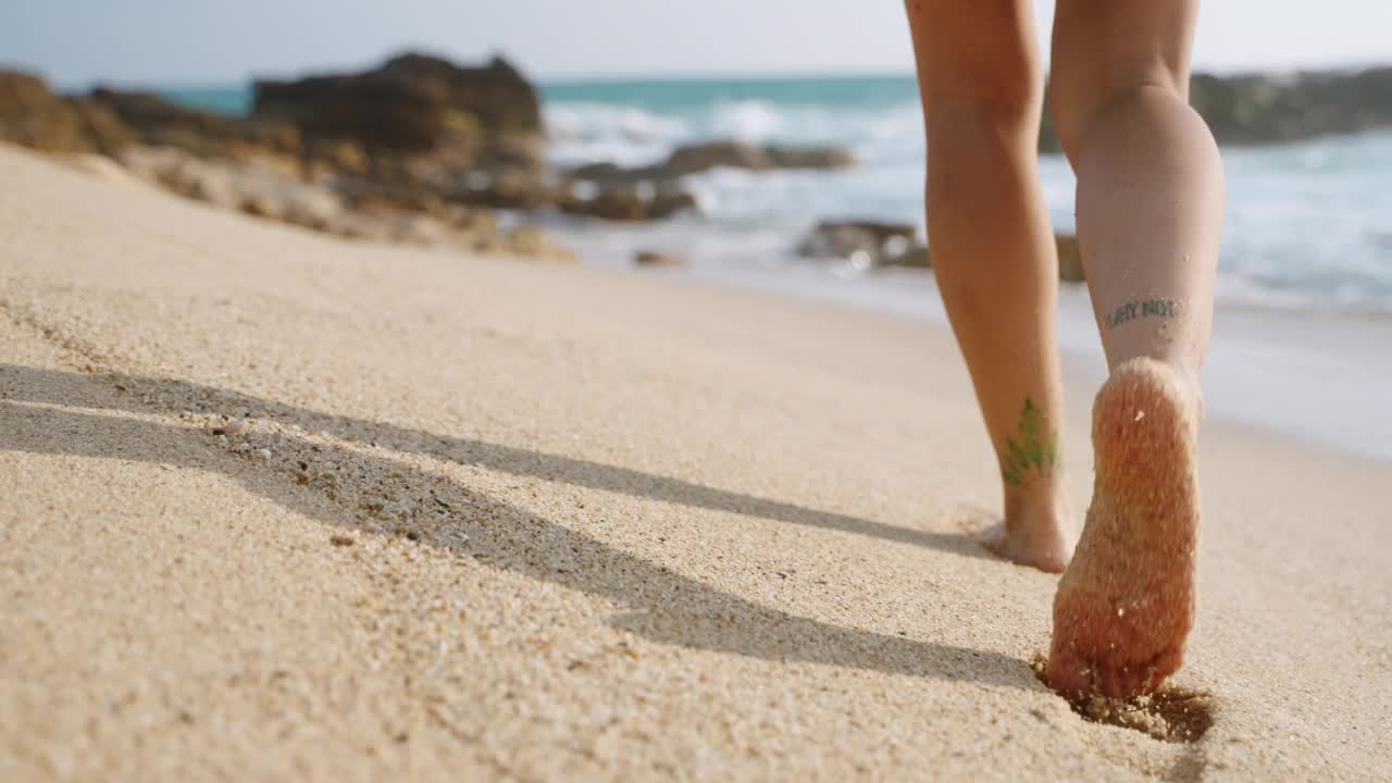 脚上有纹身的赤脚女艺术家在沙滩上拿着三脚架。创意网红漫步在水晶般的海浪中，探索美丽的海岸景观拍摄。休闲博客阳光海滩。Slomo视频下载