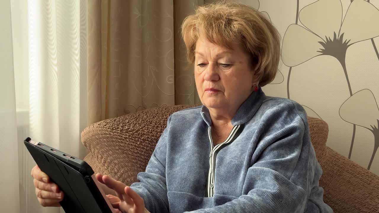 一位时髦的老妇人坐在扶手椅上，在家里用智能平板电脑查找重要信息视频下载